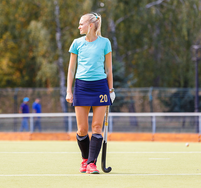 A female hockey player wearing custom sportswear—a light blue shirt, navy skirt with the number 20, black knee socks, and red trainers—stands on a pitch holding a hockey stick, looking to her left. Trees and a fence are in the background.