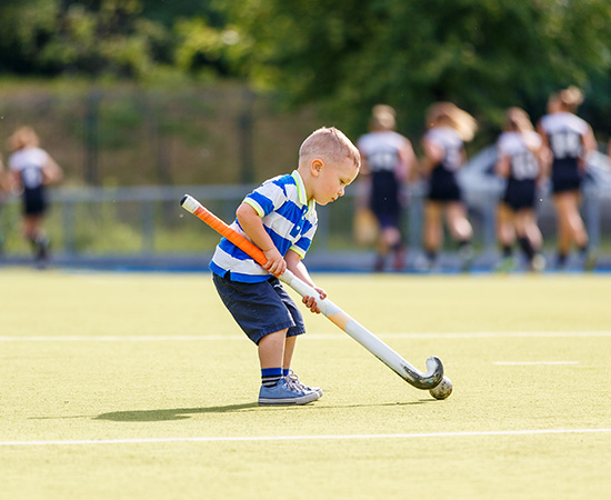 A young boy in a blue striped shirt and shorts plays hockey alone on an artificial pitch, bending down to hit the ball with his stick, while players in dark kit are blurred in the background.