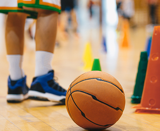 A basketball rests on a gym floor near orange and green cones. A person's legs wearing athletic shoes are visible in the background.