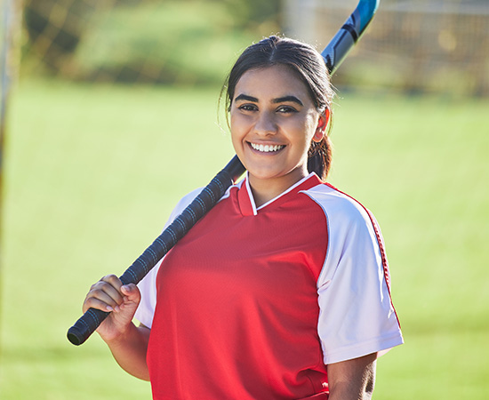 A young woman in a red and white sports jersey smiles whilst holding a hockey stick over her shoulder, standing outside on a grassy field.