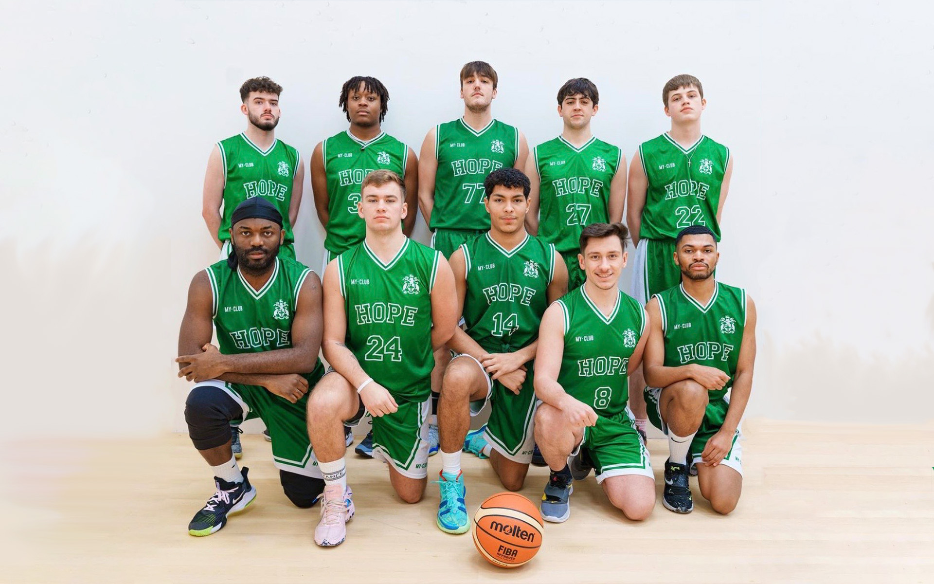 A men's basketball team wearing green jerseys poses indoors. Five players kneel in front, while six stand behind them. A basketball sits on the floor in front of the group. The word HOPE is visible on their jerseys.