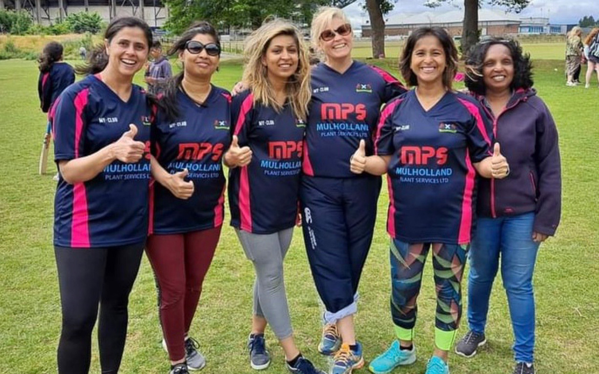 Six women stand together outdoors on grass, smiling and posing for the camera. Five wear matching navy and pink sports kits, whilst one wears a brown jacket. Some women have their thumbs up. Trees and people are visible in the background.