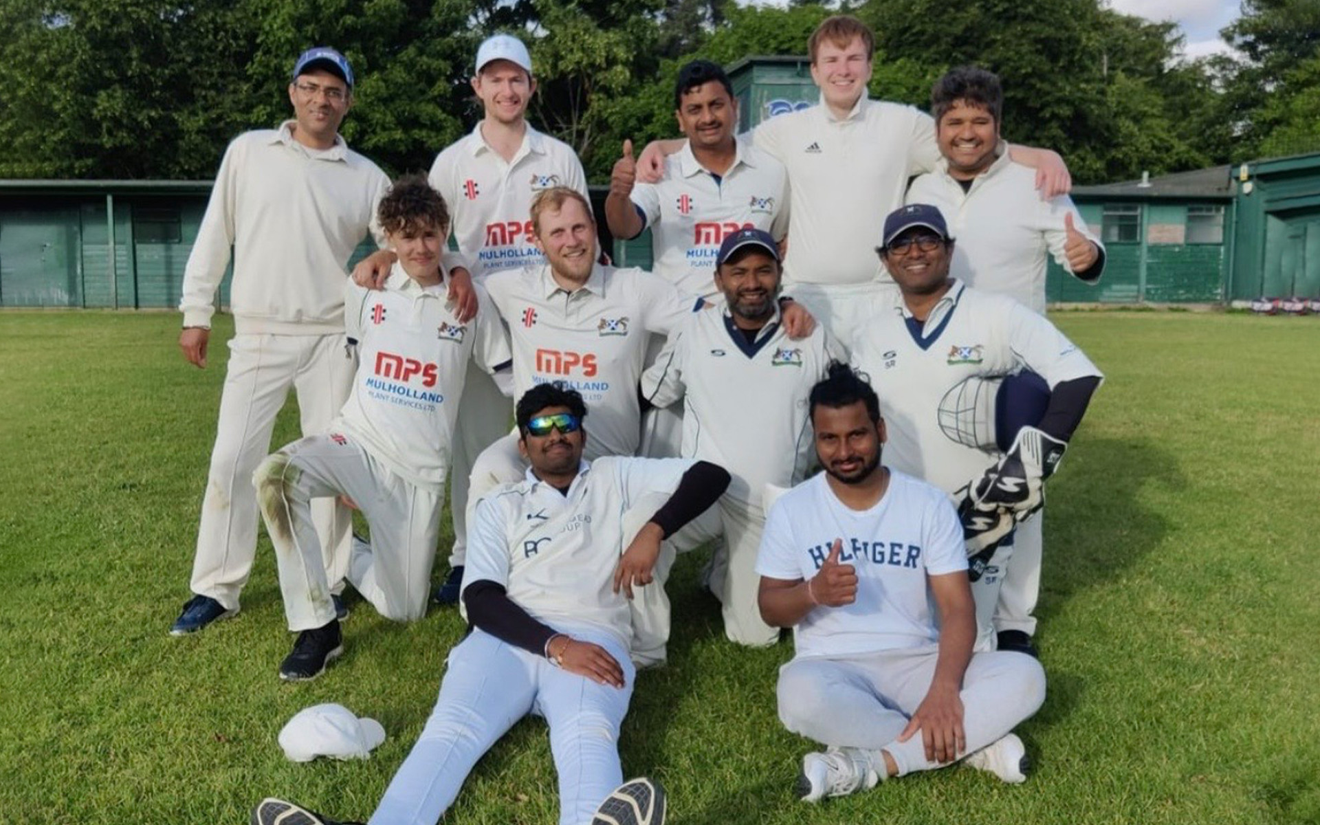 A group of eleven men in cricket kit pose together on a grass pitch, smiling at the camera. Some are standing, others are kneeling or sitting, with trees and green sheds in the background.