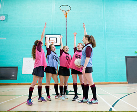 Five girls in sports uniforms practice passing a netball indoors, under a tall basketball-style hoop.