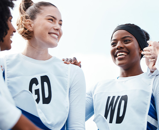 Zwei Netzballspielerinnen, die GD- und WD-Lätzchen tragen, lächeln sich im Freien an, wobei eine weitere Person an der Seite teilweise sichtbar ist.