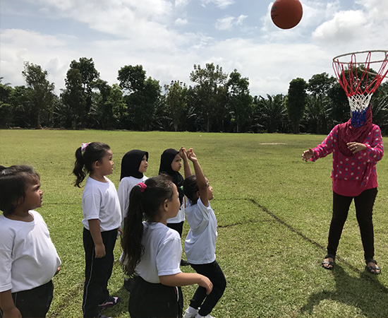 Children in white shirts play basketball outdoors with an adult supervising.