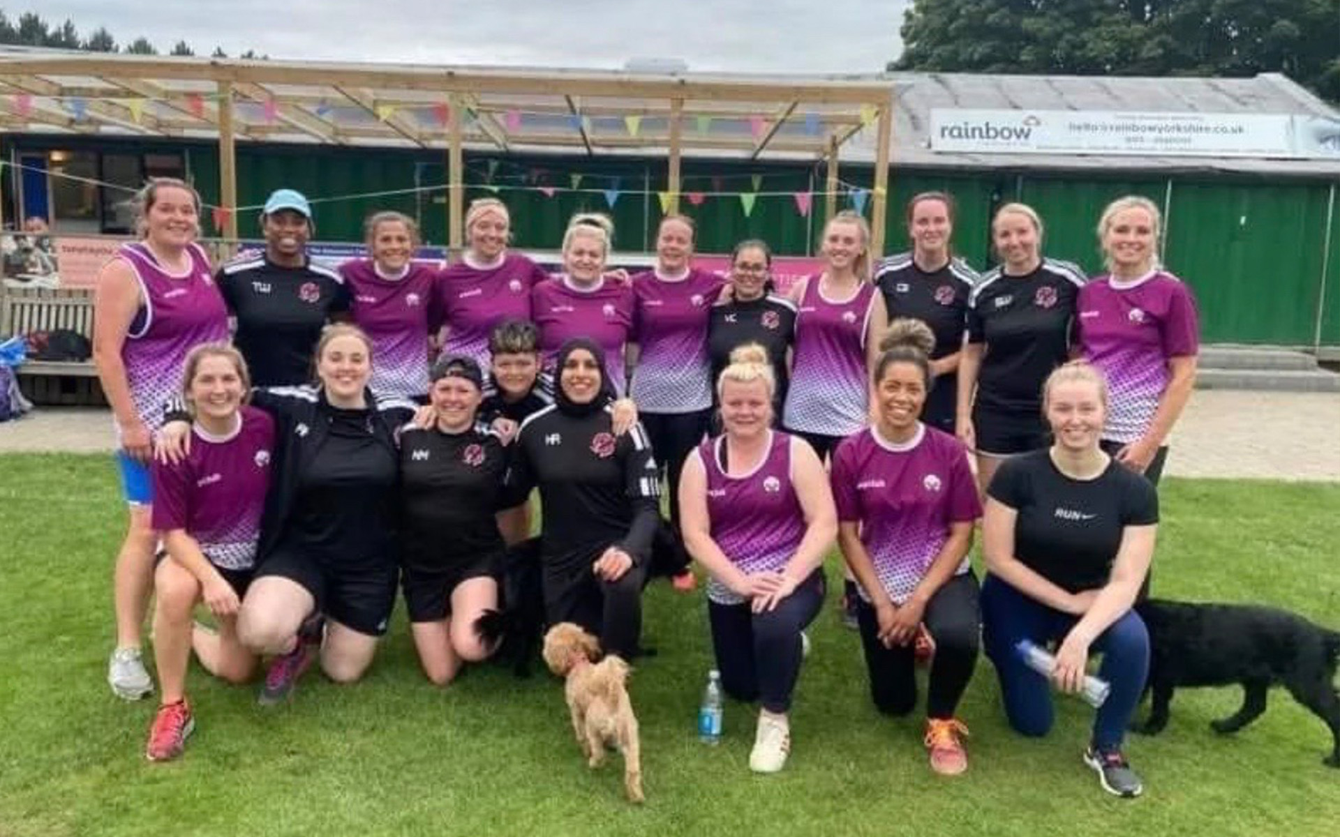 A group of women in sports kits pose together and smile on a grassy field; a small dog stands in front, and a black dog is partly visible on the right. A building and colourful bunting are in the background.
