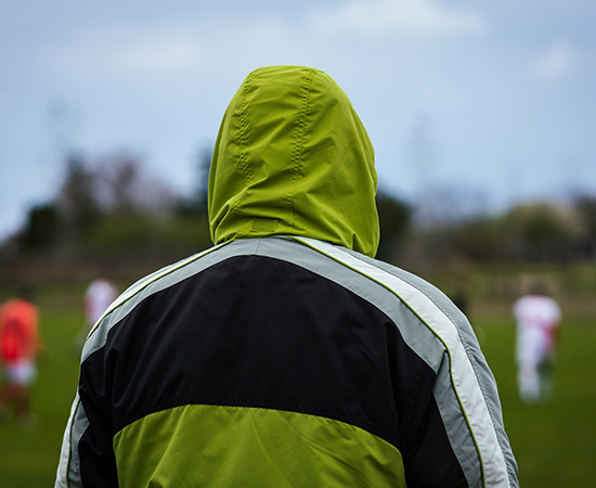 Une personne portant une veste à capuche verte se tient à l'extérieur, face à un terrain de sport avec des joueurs flous en arrière-plan.