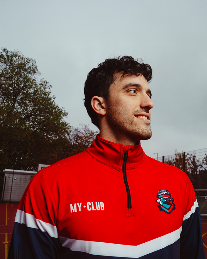 Une personne portant un maillot de sport rouge se tient à l'extérieur et sourit. Le maillot porte l'inscription "MY CLUB" et un logo. Des arbres et un ciel nuageux sont à l'arrière-plan.