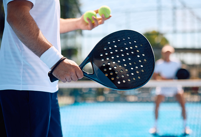 Nahaufnahme einer Person, die einen Padel-Schläger und zwei Bälle auf einem Platz im Freien hält, mit einem anderen Spieler im Hintergrund, der zum Spiel bereit ist. Beide Spieler tragen weiße Sportkleidung.