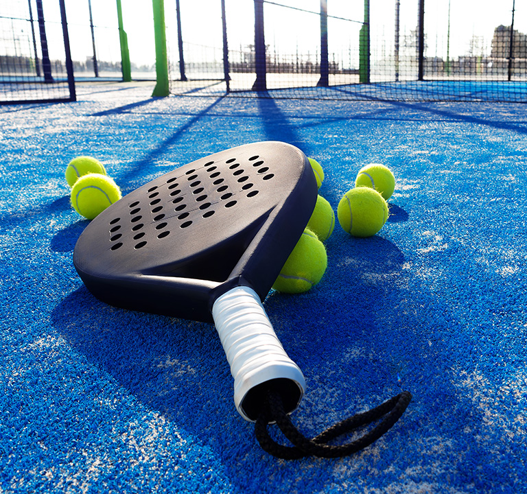 Ein schwarzer Padel-Schläger mit weißem Griff liegt auf einem hellblauen Spielfeld, umgeben von mehreren gelben Tennisbällen, mit einem Netz und einer Umzäunung im Hintergrund.