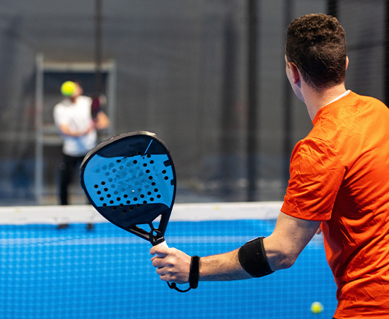 Ein Mann in einem orangefarbenen Hemd hält einen Padel-Schläger und bereitet sich darauf vor, einen Ball zu schlagen, während ein anderer Spieler im Hintergrund auf einem blauen Padel-Platz steht.
