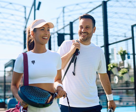 Eine lächelnde Frau und ein lächelnder Mann in Sportkleidung gehen gemeinsam auf einem Padel-Platz im Freien, jeder trägt einen Schläger. Der Hintergrund zeigt einen eingezäunten Platz und einen klaren Himmel, der auf einen sonnigen Tag hindeutet.