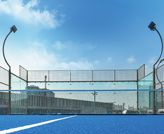 Ansicht eines leeren Padel-Tennisplatzes mit blauem Belag, weißen Spielfeldlinien, einem Netz im Vordergrund und hohen Lichtern unter einem klaren blauen Himmel.