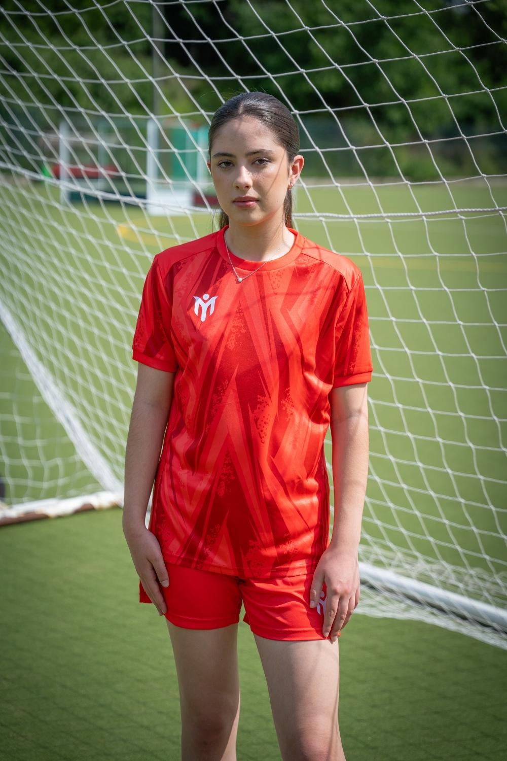 Eine junge Frau in einem roten Fußballtrikot steht vor einem Fußballtor auf einem grünen Spielfeld und schaut mit neutralem Blick in die Kamera.