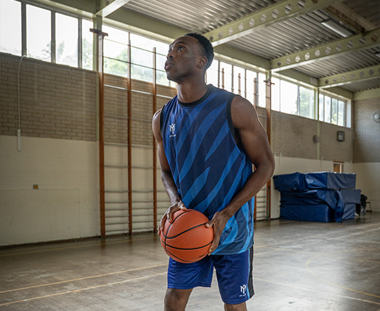 A basketball player in a blue sports kit stands in a gymnasium, holding a basketball and looking upwards, appearing focused and ready to take a shot. Gym equipment and stacked mats are visible in the background.