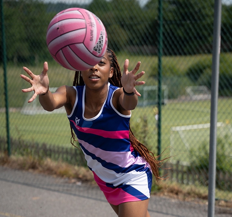 A young woman in a blue, pink, and white netball kit reaches out to catch a pink netball during an outdoor match on a sunny day, with a green fence and grassy pitch in the background.
