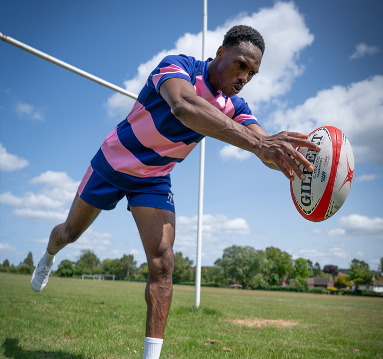 A rugby player in a blue and pink kit dives to score a try, extending the ball towards the ground near the goalposts on a grassy pitch under a partly cloudy sky.