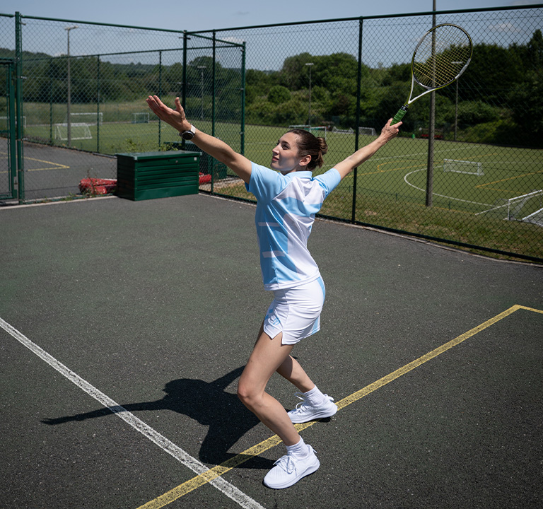 Une femme en tenue de sport se tient sur un court de tennis, s'apprêtant à servir avec une raquette levée. Elle est concentrée, le bras tendu vers le haut, et le soleil brille sur le court extérieur.