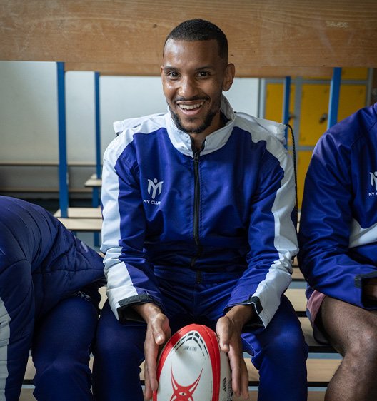 Un homme en survêtement bleu et blanc est assis sur un banc dans un vestiaire, souriant et tenant un ballon de rugby. Deux coéquipiers sont assis à ses côtés, partiellement visibles.