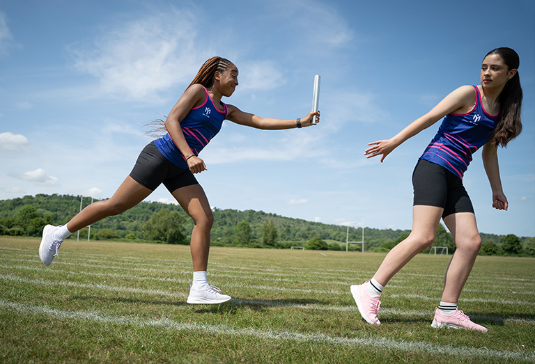 Deux athlètes féminines en tenue bleue et noire courent sur une piste herbeuse, l'une tendant le bras pour passer un bâton de relais à l'autre, sous un ciel bleu clair et des collines vertes en arrière-plan.