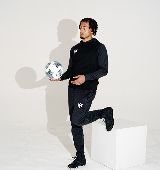 Une personne en tenue de sport noire pose avec une jambe sur un cube blanc, tenant un ballon de football dans une main, sur un fond blanc uni.