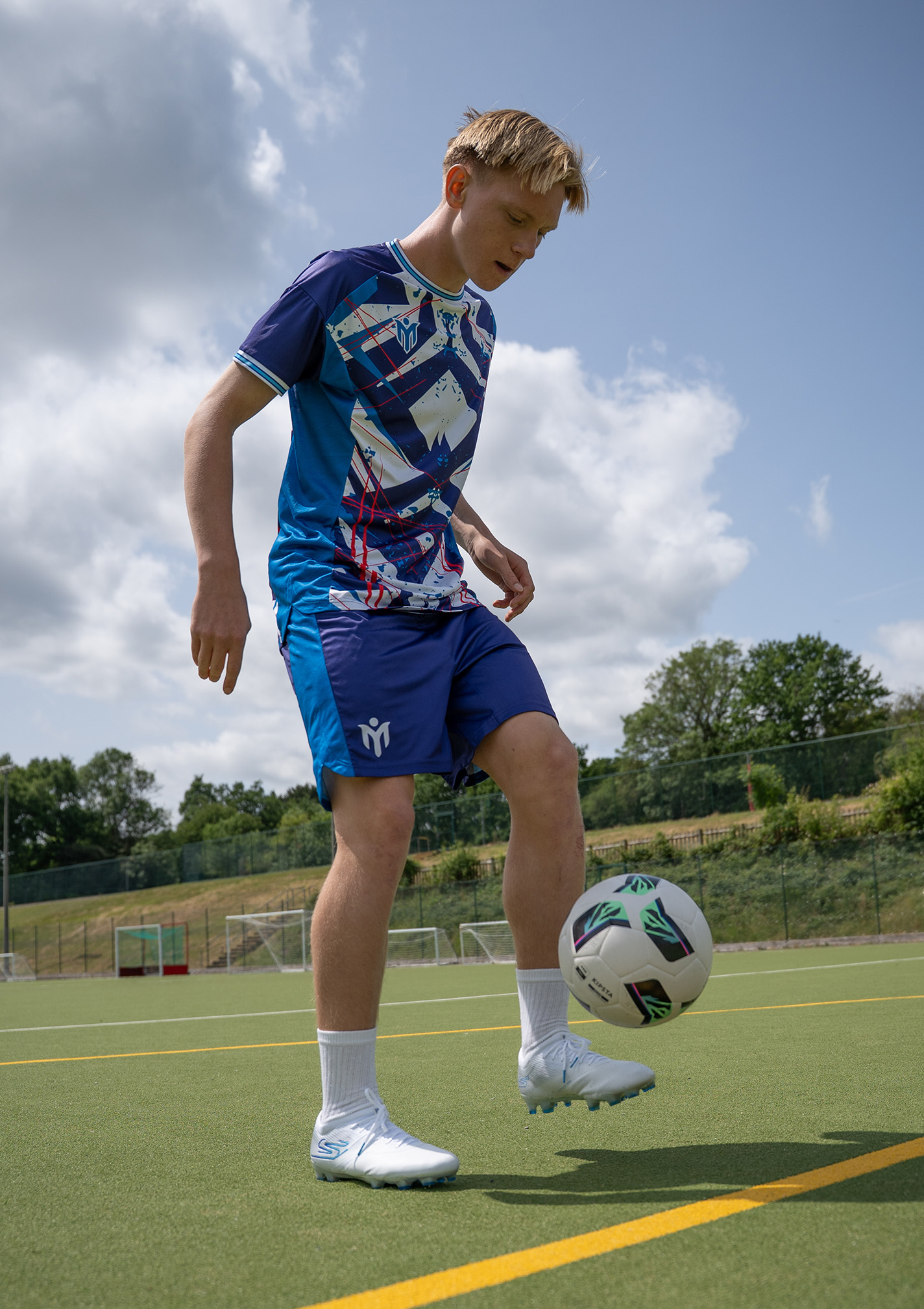 A teenage boy in a blue sports kit is juggling a football on an outdoor pitch under a partly cloudy sky. Trees and football goals are visible in the background.