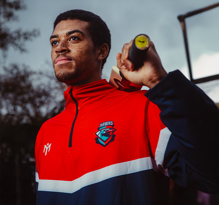 A person in a red and blue HAWKS sports jacket holds a baseball bat over their shoulder, looking confidently into the distance with an overcast sky in the background.