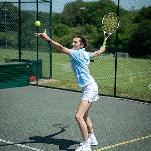 Una mujer vestida con un conjunto azul claro y blanco se dispone a sacar una pelota de tenis en una pista al aire libre, sosteniendo la raqueta por encima de la cabeza con una mano y lanzando la pelota al aire con la otra. Al fondo se ven unos árboles y una valla.