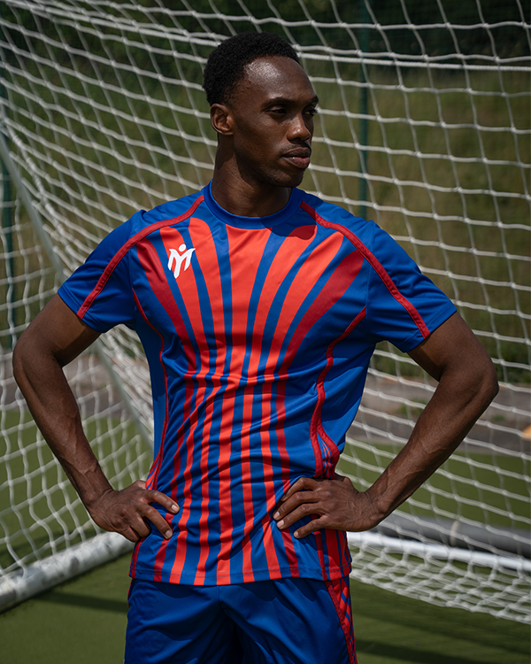 Ein Fußballspieler in einem blau-rot gestreiften Hemd steht mit den Händen in den Hüften vor einem Tornetz auf einem grünen Fußballplatz.