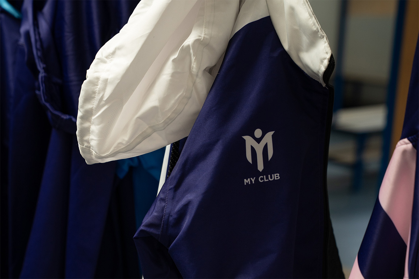 Primer plano de una chaqueta deportiva azul y blanca colgada de una barandilla, con un logotipo con una persona estilizada y el texto MY CLUB impreso en la espalda. Otras chaquetas cuelgan junto a ella en el fondo.