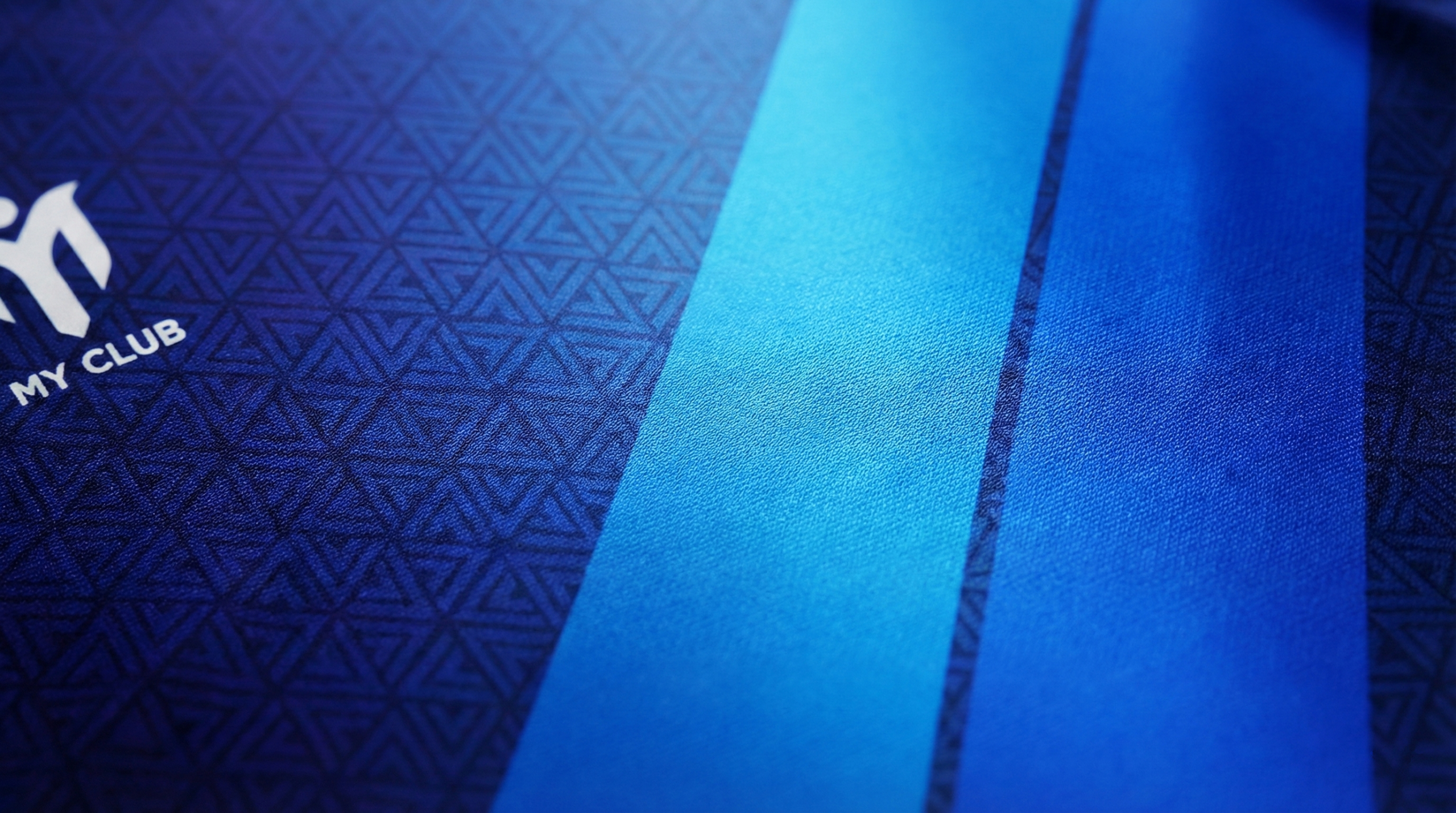 Primer plano de una camiseta de fútbol azul personalizada con un motivo geométrico triangular y dos rayas verticales azul claro. En el lado izquierdo se ve un logotipo parcial blanco con las palabras MY CLUB.