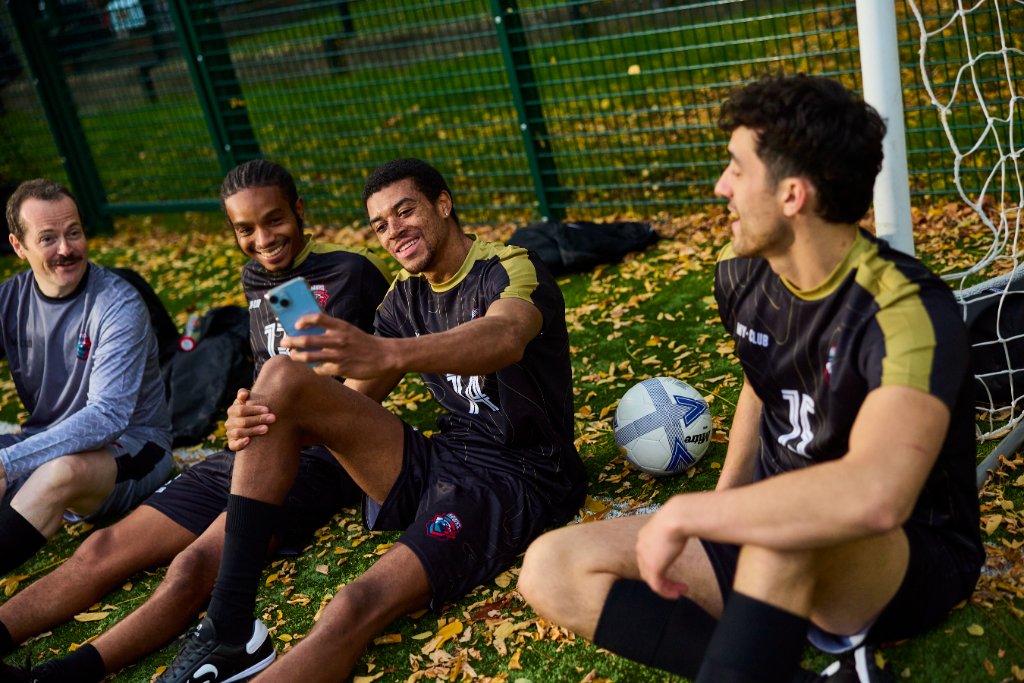 Quatre footballeurs, vêtus de tenues de football personnalisées, sont assis sur la pelouse près d'un but ; ils sourient et rient tandis que l'un d'entre eux prend un selfie. Des feuilles d'automne jonchent le sol et une clôture métallique est visible à l'arrière-plan.