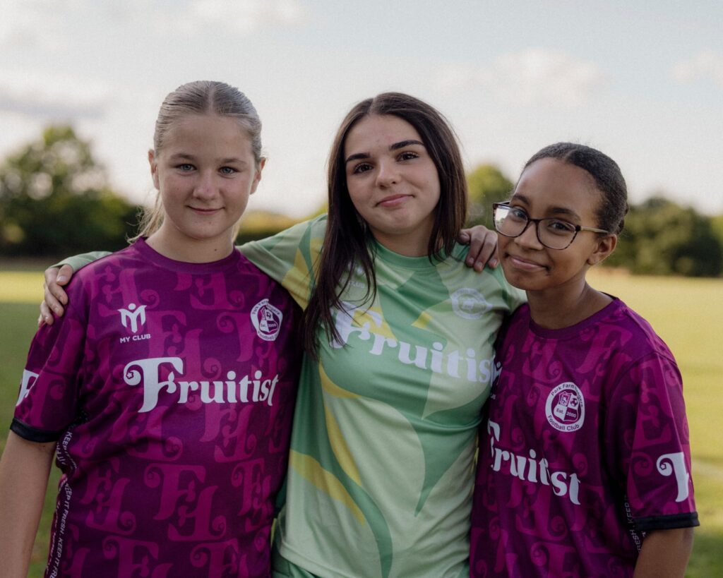 Tres chicas jóvenes están juntas en un campo de hierba, sonriendo a la cámara. Dos llevan camisetas deportivas moradas a juego que dicen Fruitist, y una lleva una camiseta verde con el mismo logotipo. Al fondo se ven árboles y el cielo.