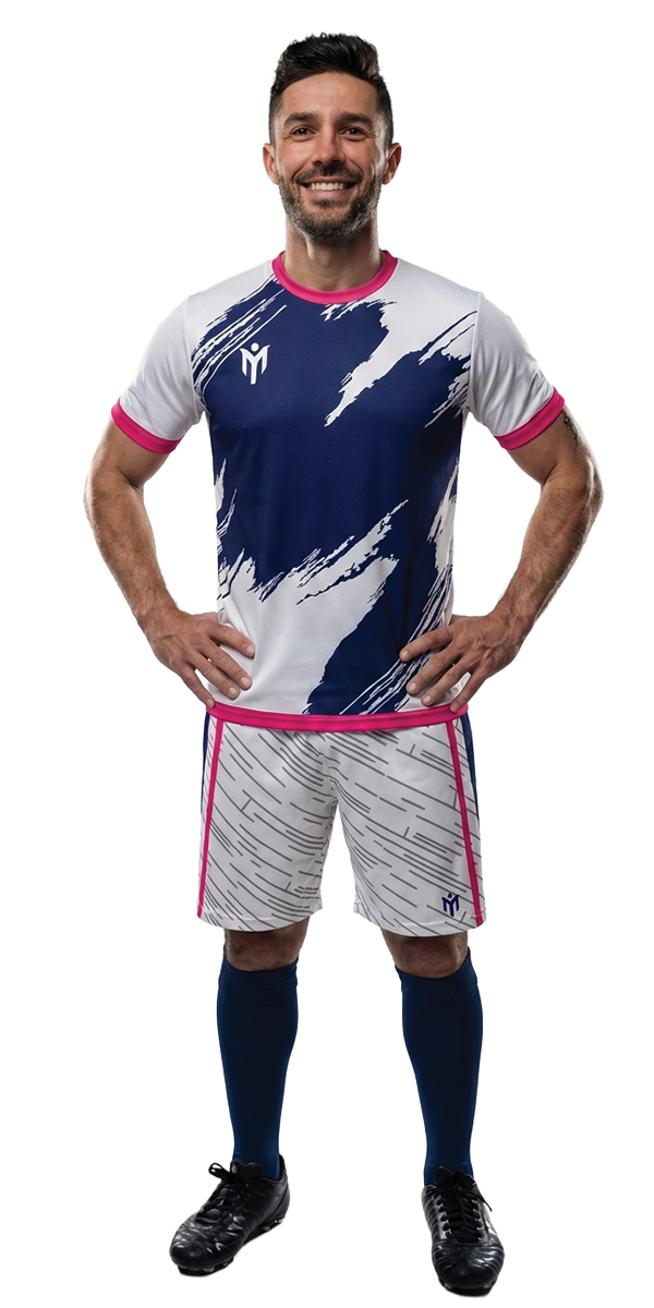 Ein lächelnder Mann steht mit den Händen in den Hüften, trägt ein weißes und marineblaues Fußballtrikot mit rosa Akzenten, marineblaue Socken und schwarze Stiefel vor einem schlichten weißen Hintergrund.