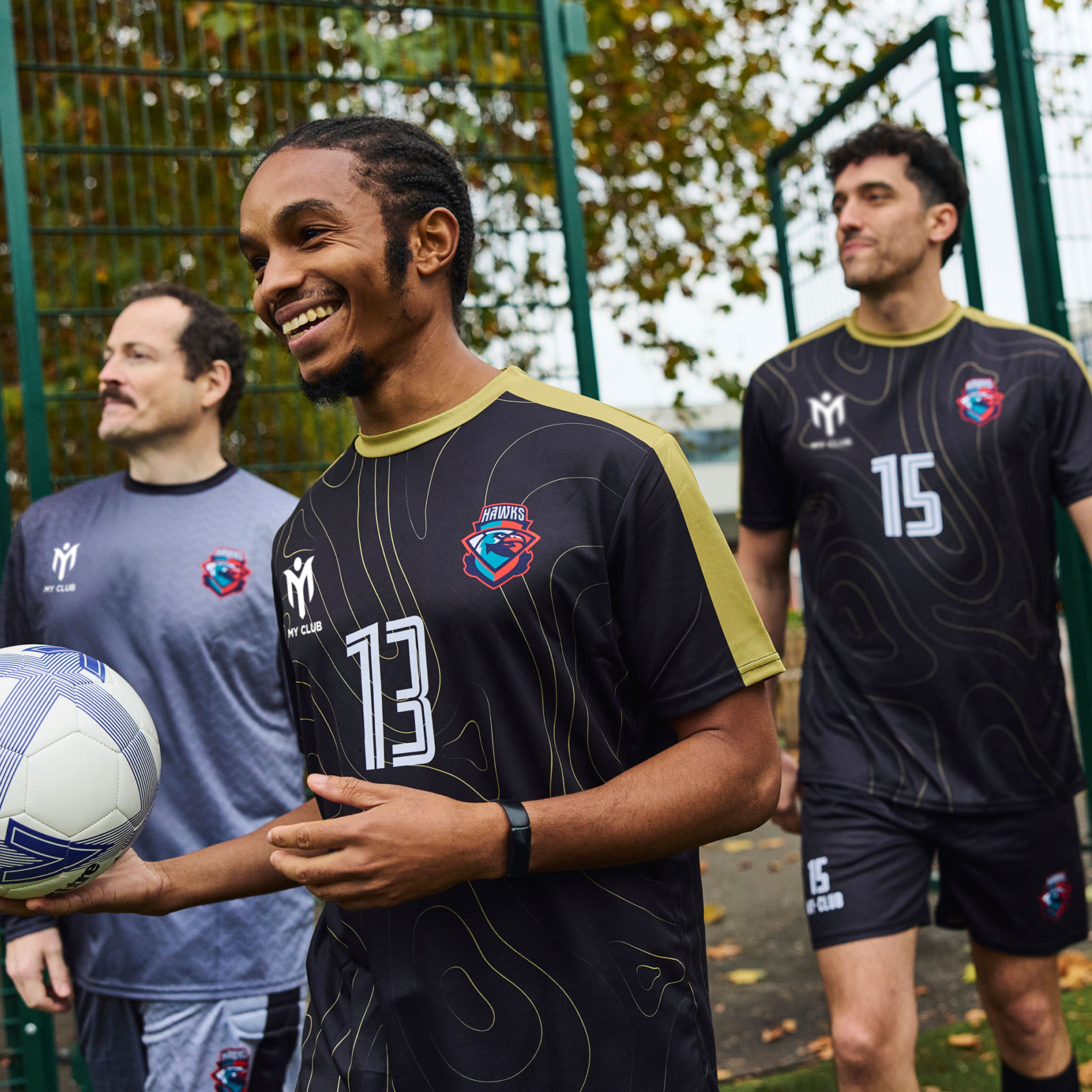 Three men wearing matching custom football kits stand outside by a green metal fence. One holds a football. They are smiling and appear ready for a match, with trees and outdoor scenery in the background.