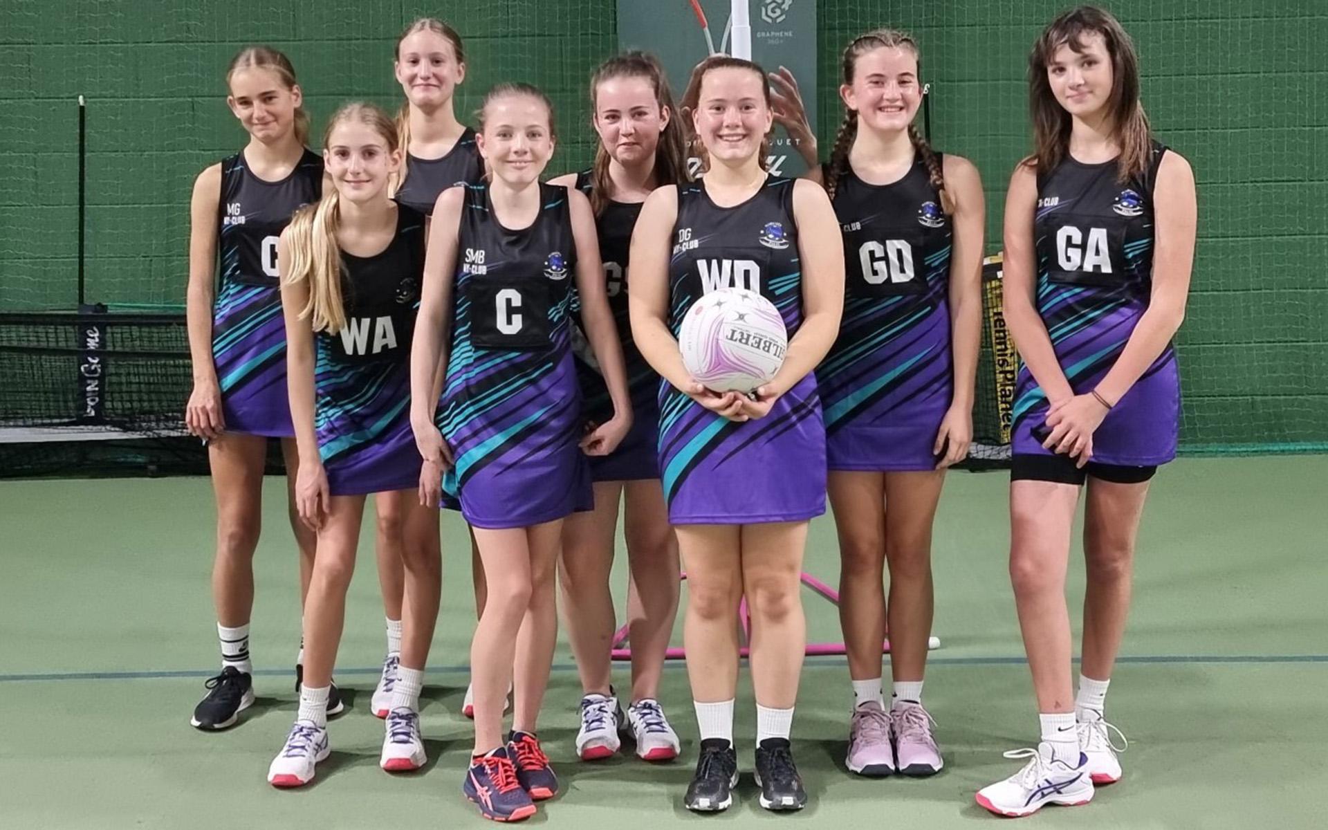 Eight girls wearing matching netball kits pose for a team photo on an indoor court. One girl at the front holds a netball, and all are smiling, standing in two rows against a green netted background.