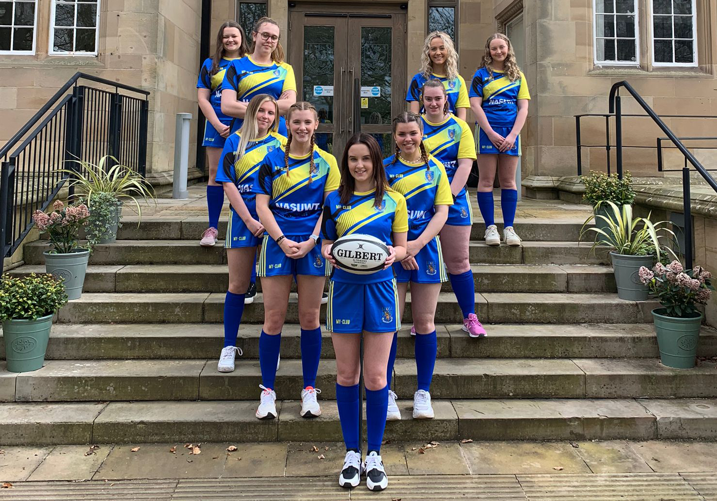 Eight women in blue and yellow rugby kits stand in two rows on stone steps outside a building. The front row holds a rugby ball, and potted plants line the steps. All are smiling at the camera.