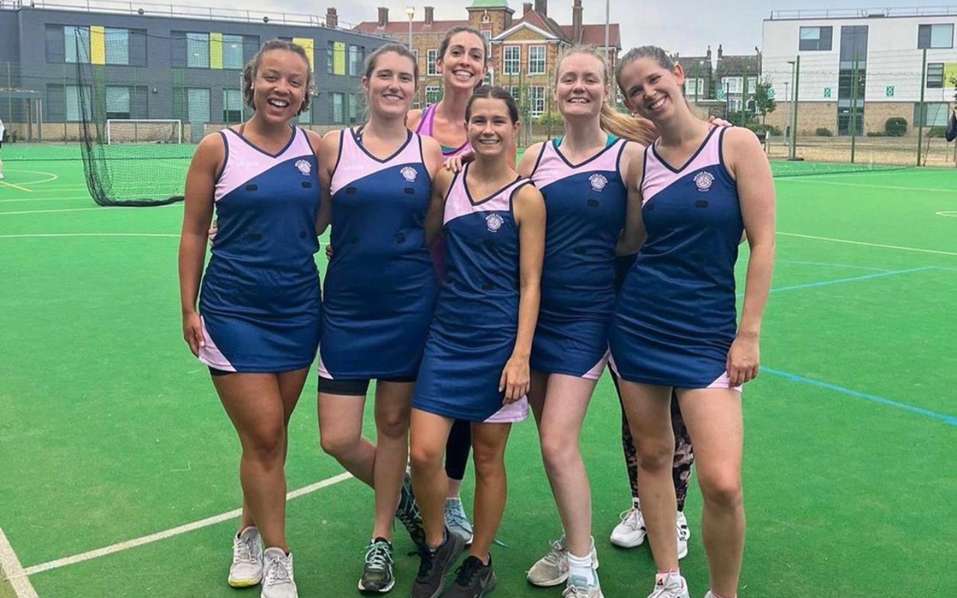 Sei donne in divisa da netball blu e rosa sono in posa insieme su un campo all'aperto, sorridendo alla fotocamera con le braccia intrecciate. Sullo sfondo si vedono alcuni edifici e una rete.