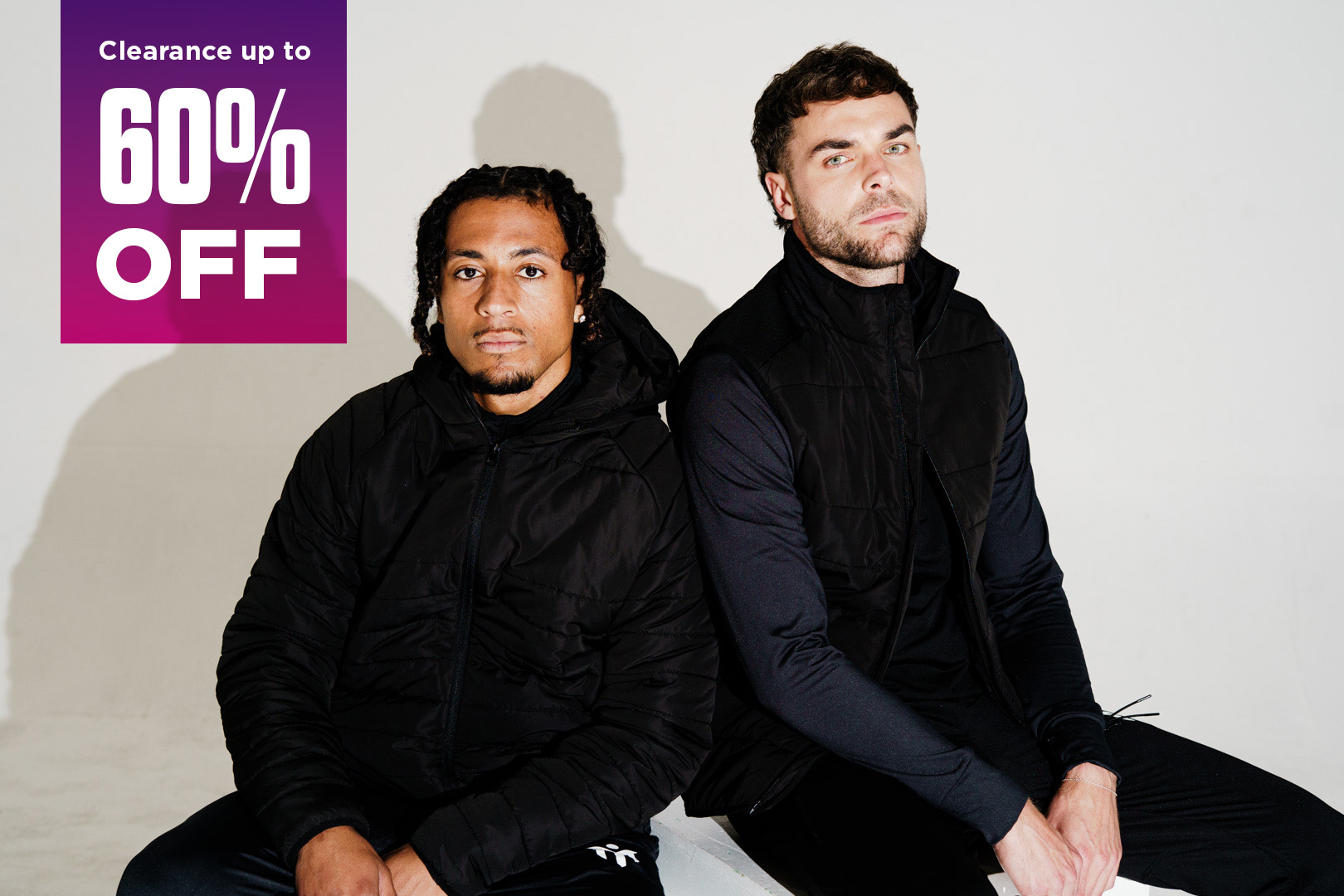 Two men in black sportswear sit on a white bench against a plain background. A purple banner in the top left corner reads Clearance up to 60% OFF in bold white and pink text.