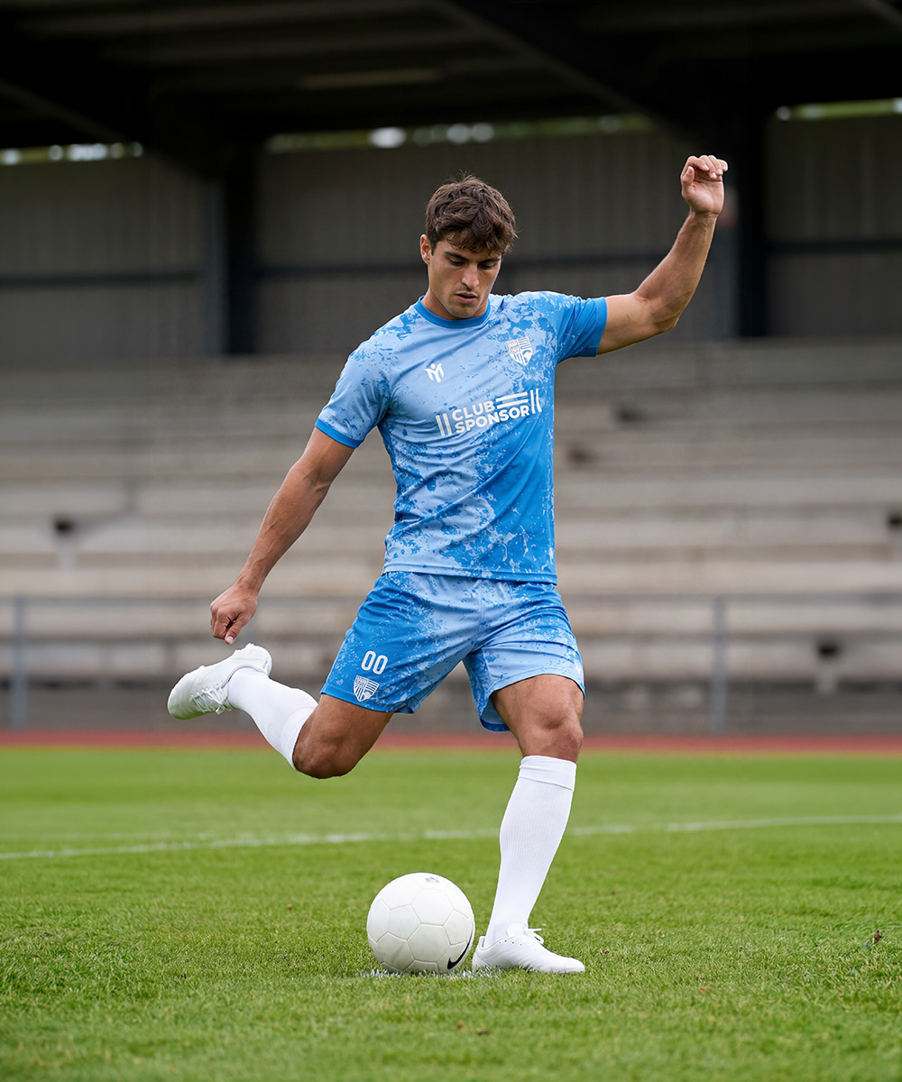 Un calciatore, con indosso una maglia personalizzata nei colori blu e bianco, sta per calciare un pallone bianco su un campo in erba, con le tribune vuote sullo sfondo.
