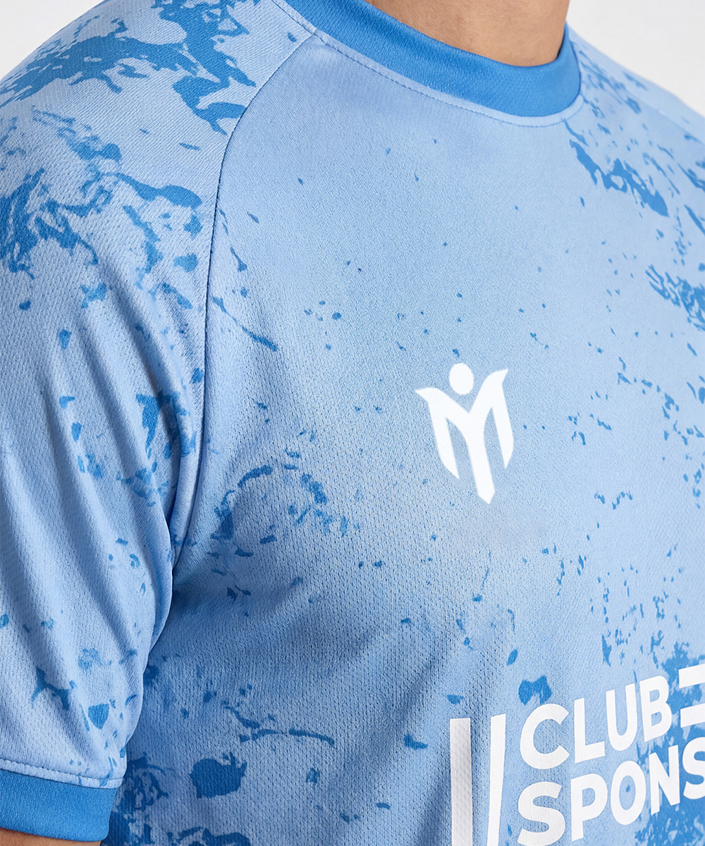 Un primo piano di una persona che indossa una maglia sportiva azzurra con motivi a spruzzi blu scuro, caratterizzata da un logo astratto bianco sul petto e da una scritta parzialmente visibile che recita “CLUB SPONS”: perfetta per gli appassionati di maglie da calcio personalizzate.