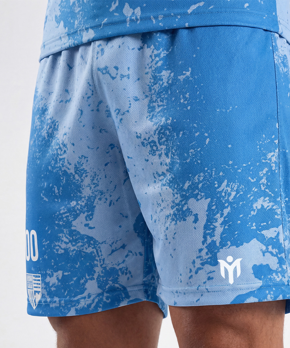Un primo piano di una persona che indossa pantaloncini da calcio blu con un motivo astratto bianco, con loghi e il numero 00 sulla gamba sinistra.