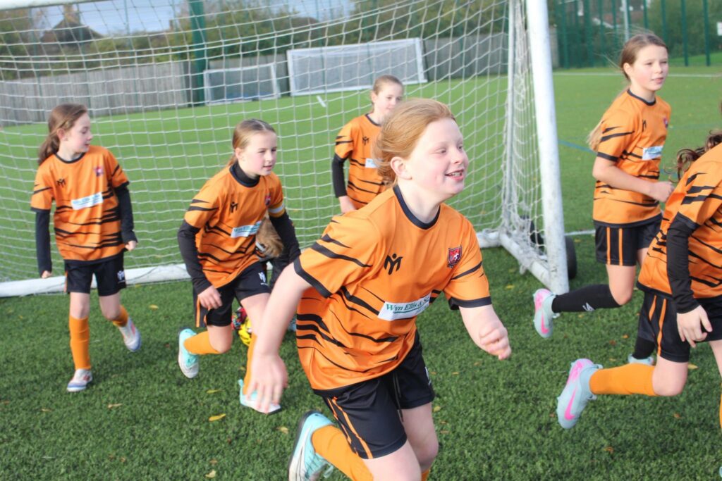 A group of young girls in orange and black football kits run joyfully in front of a goal on a green football pitch during the day.