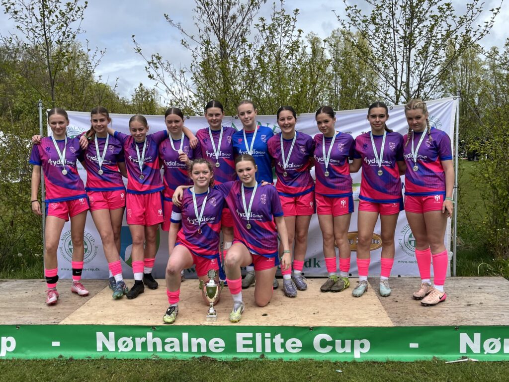 A girls’ football team in matching pink and purple sustainable sportswear stands on a podium with medals, posing behind a trophy at the Nørhalne Elite Cup. Green trees and a banner form the backdrop.
