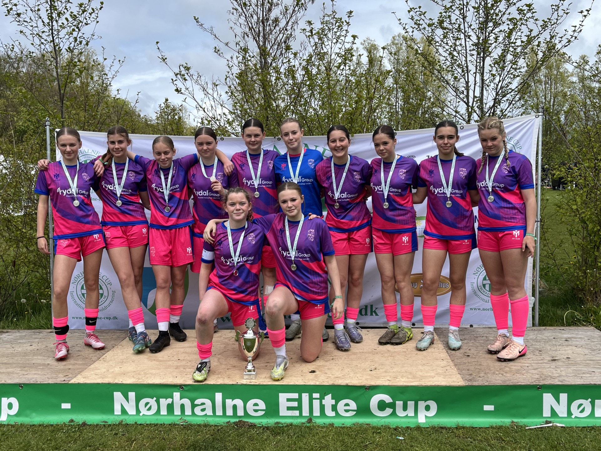 A girls&rsquo; football team in matching pink and purple sustainable sportswear stands on a podium with medals, posing behind a trophy at the N&oslash;rhalne Elite Cup. Green trees and a banner form the backdrop.