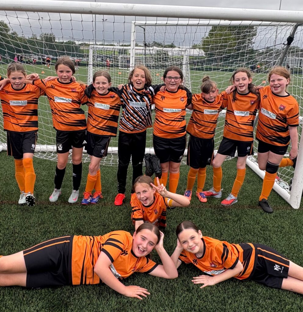 Una squadra di calcio femminile in tenuta arancione e nera posa davanti a una porta su un campo erboso, mostrando come le società sportive di base aiutino i giovani atleti a crescere. Otto ragazze stanno a braccetto, mentre tre sono sdraiate sul manto erboso, sorridendo e facendo segni di pace.