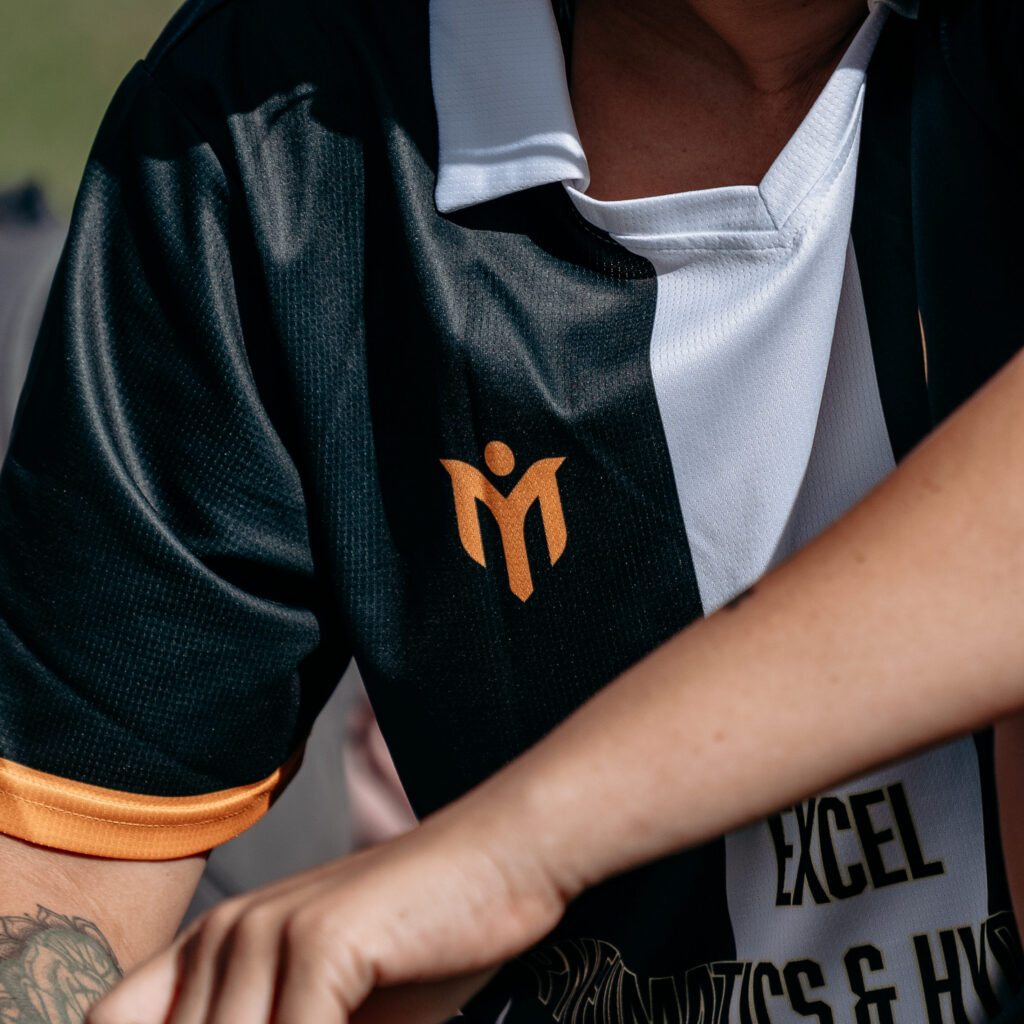 Eine Person, die ein schwarz-weißes Sporttrikot mit orangefarbenen Akzenten und einem Logo trägt, sitzt im Freien und hält eine Wasserflasche in der Hand. Auf dem Arm der Person ist ein Tattoo zu sehen, und sie schaut zur Seite.