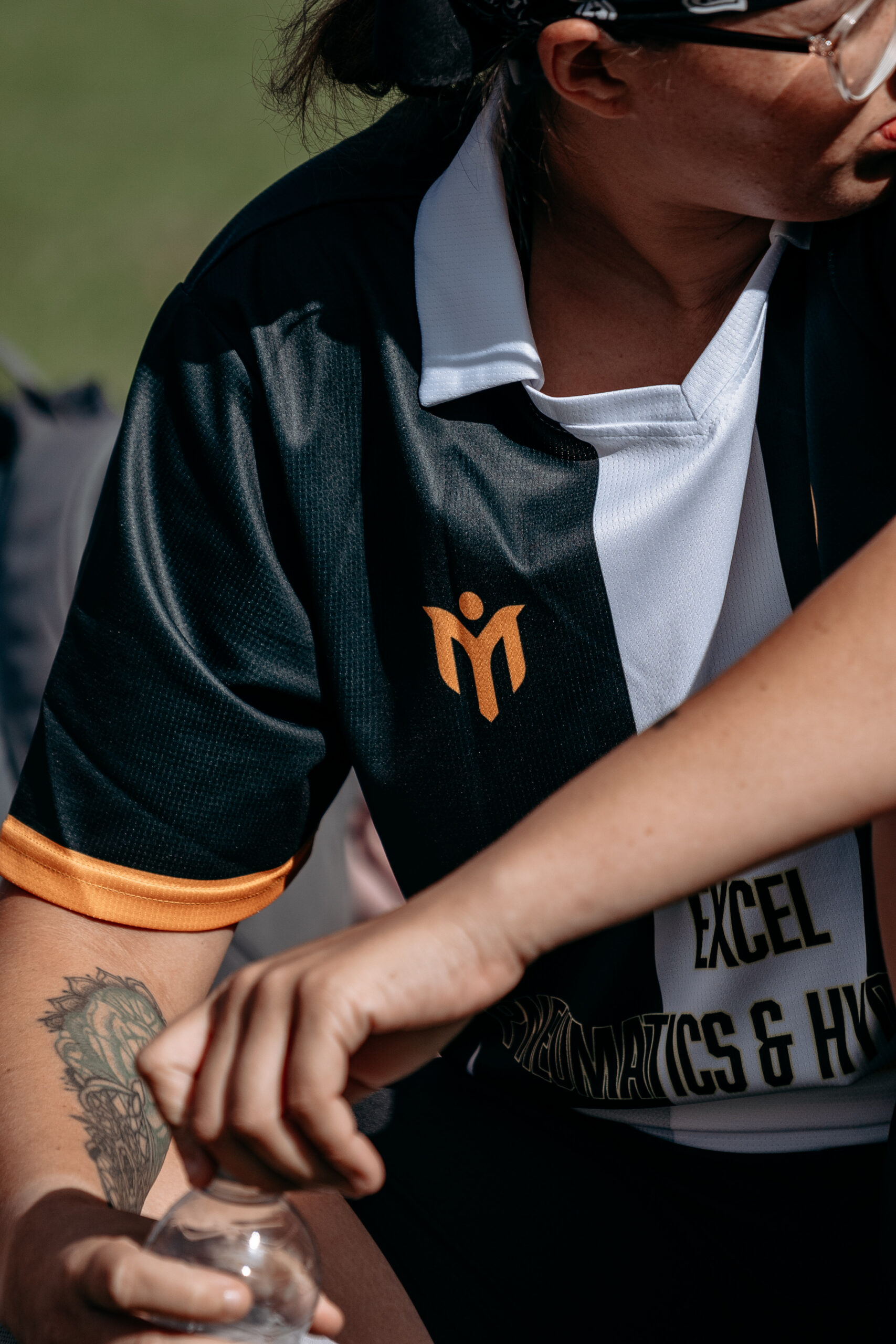 Persona che indossa una maglia sportiva bianca e nera con dettagli arancioni e un logo, seduta all’aperto, con in mano una bottiglia d’acqua. Ha un tatuaggio sul braccio e guarda di lato.