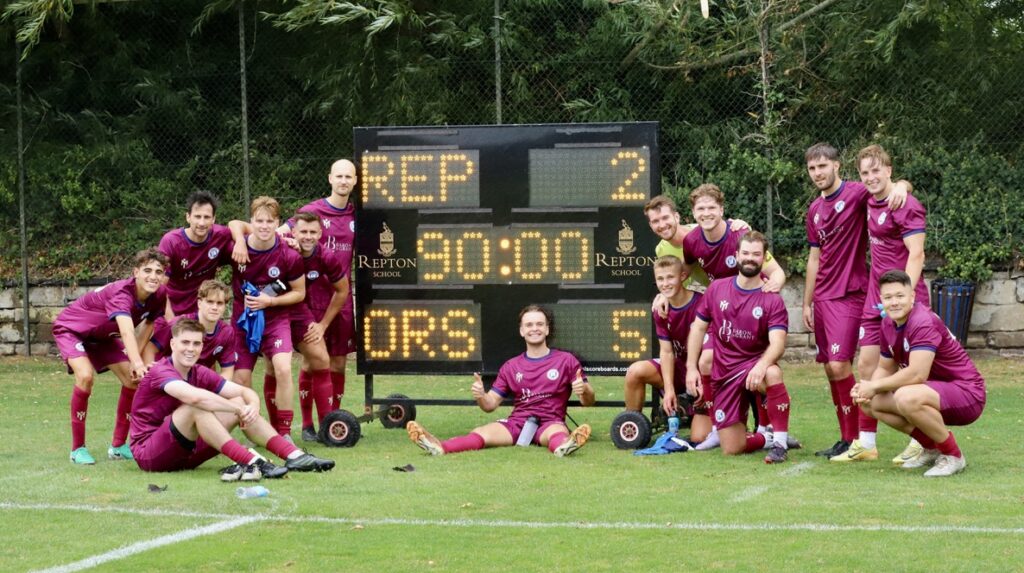 Un equipo de fútbol con equipaciones granates posa alegremente delante de un marcador que indica «REP 2 - ORS 5» tras un partido disputado en un campo de césped, con árboles y una valla al fondo.